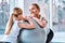 Beautiful young mother sits together with her daughter on the floor near fitball and they keep dumbbells in their hands, at the