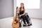 Beautiful young musician with guitar sitting in wheelchair indoors