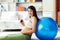 Beautiful young pregnant asian woman holding apple and lean against exercise ball in living room at house with smiley face and