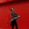 Beautiful young slim attractive woman with sunglasses in motion dresses a green fashionable coat near the red metal wall.