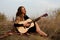 beautiful young smiling woman playing melody on acoustic guitar while sitting in field among tall dried grass.