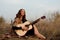 beautiful young smiling woman playing melody on acoustic guitar while sitting in field among tall dried grass.