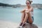 Beautiful young stylish slim blonde in short and white top in sunglasses sits on the seashore against the blue sky, fashion and