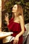 A beautiful young woman in an asymmetrical red-burgundy cocktail dress on one shoulder sits in a restaurant with glass