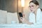 Beautiful young woman in bathrobe having rest on comfortable bed room after spa procedures. She is typing message on smartphone