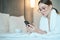 Beautiful young woman in bathrobe having rest on comfortable bed room after spa procedures. She is typing message on smartphone