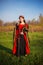 A beautiful, young woman in a black and red medieval dress with a crown on her head stands in a field with a sword in her hands. C