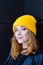 Beautiful young woman with blonde hair and blue eyes in a yellow knitting hat on a background of black wooden wall.