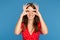 Beautiful young woman with bright smile in red dress having fun making glasses shape with hands over blue background.