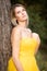 Beautiful young woman with clean perfect skin wearing a classy yellow evening dress, leaning against a tree in the