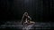 Beautiful young woman with dark hair in dove pose under the streams of rain. The studio light reflects on the water and