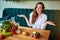 Beautiful young woman decides eating hamburger or vegetables  in kitchen. Cheap junk food vs healthy diet