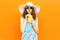 Beautiful young woman drinking juice in summer straw hat, colorful striped dress on orange wall