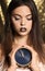 Beautiful young woman with extravagant makeup holding clock against defocused lights