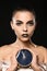 Beautiful young woman with extravagant makeup holding clock on black background