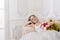 Beautiful young woman with flowers dressed in a white full wide-skirted dress sitting on the sofa