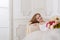 Beautiful young woman with flowers dressed in a white full wide-skirted dress sitting on the sofa