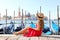 Beautiful young woman with hat sitting on the edge of Venetian Lagoon looking at stunning panoramic view of Venice, Italy
