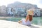 Beautiful young woman with hat sitting looks at stunning panoramic village of Cefalu on Sicily Island, Italy