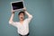 Beautiful young woman holding netbook computer looking up and saying wow wearing white shirt isolated on blue background