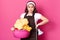 Beautiful young woman holds pink basin with laundry and iron, looking at camera while standing isolated over rose background,