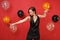 Beautiful young woman in little black dress celebrating dancing spreading hands pointing finger on bright red background