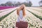 Beautiful young woman with long red hair wearing in white dress standing with luggage on colorful tulip field