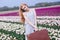 Beautiful young woman with long red hair wearing in white dress standing with luggage on colorful tulip field