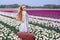 Beautiful young woman with long red hair wearing in white dress standing with luggage on colorful tulip field
