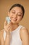 Beautiful young woman looking happy while using silicone facial cleansing brush, posing isolated over beige background