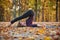 Beautiful young woman practices yoga asana Halasana Plough pose on the wooden deck in the autumn park.