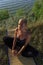 Beautiful young woman practising yoga on mat outdoors at river bank on grass