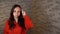 A beautiful young woman in a red hoodie twists at her temple on a gray brick background.