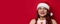 Beautiful young woman with red santa hat with pompom expressing happy emotions and holding present box on red wall background