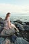 Beautiful young woman relaxing on ocean coast, romantic beauty portrait. Pretty girl sitting on rocks and dreaming