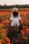 Beautiful young woman in short white dress and straw hat in field with poppies in evening at sunset and holds poppy in hand,