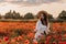 Beautiful young woman in short white dress and straw hat in field with poppies in evening at sunset and holds poppy in hand,