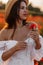 Beautiful young woman in short white dress and straw hat in field with poppies in evening at sunset and holds poppy in hand,