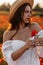 Beautiful young woman in short white dress and straw hat in field with poppies in evening at sunset and holds poppy in hand,