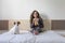 beautiful young woman sitting on bed with her cute small dog besides. Home, indoors and lifestyle. She is drinking orange juice.