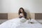 Beautiful young woman sitting on bed with her cute small dog besides. She is working on tablet and smiling. Breakfast at home,