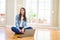 Beautiful young woman sitting on the floor with crossed legs using laptop looking away to side with smile on face, natural