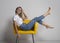 Beautiful young woman is sitting or lying on yellow chair with legs up and looking arrogantly at camera