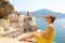 Beautiful young woman sitting on wall with panoramic view of Atrani village on Amalfi Coast, Italy