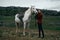 Beautiful young woman standing in the mountains and holding a white horse, nature