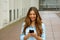 Beautiful young woman is using an app in her smartphone device to send a text message while standing in courtyard of office blocks