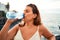 Beautiful young woman walking on beach promenade enjoying ocean view drinking fresh bottle of water