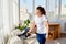 Beautiful young woman in white shirt and jeans cleaning sofa with vacuum cleaner in living room, copy space. Housework.