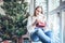 A beautiful young woman in a white sweater dress unpacks New Year`s presents near New Year tree