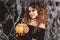 Beautifull woman in a witch`s costume is holding pumpkin in front of black wall with spider net